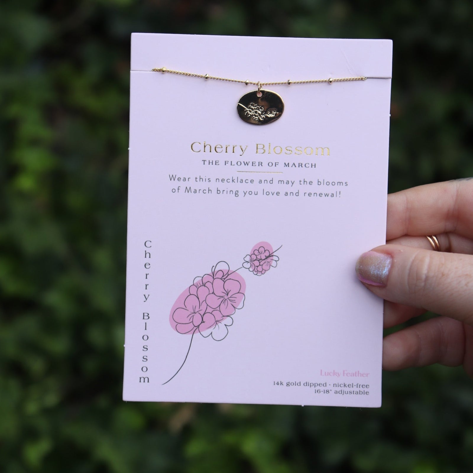 Someone holding the cherry blossom 14k Gold Dipped Nickel-Free Birth Month Flower Necklace in front of greenery.
