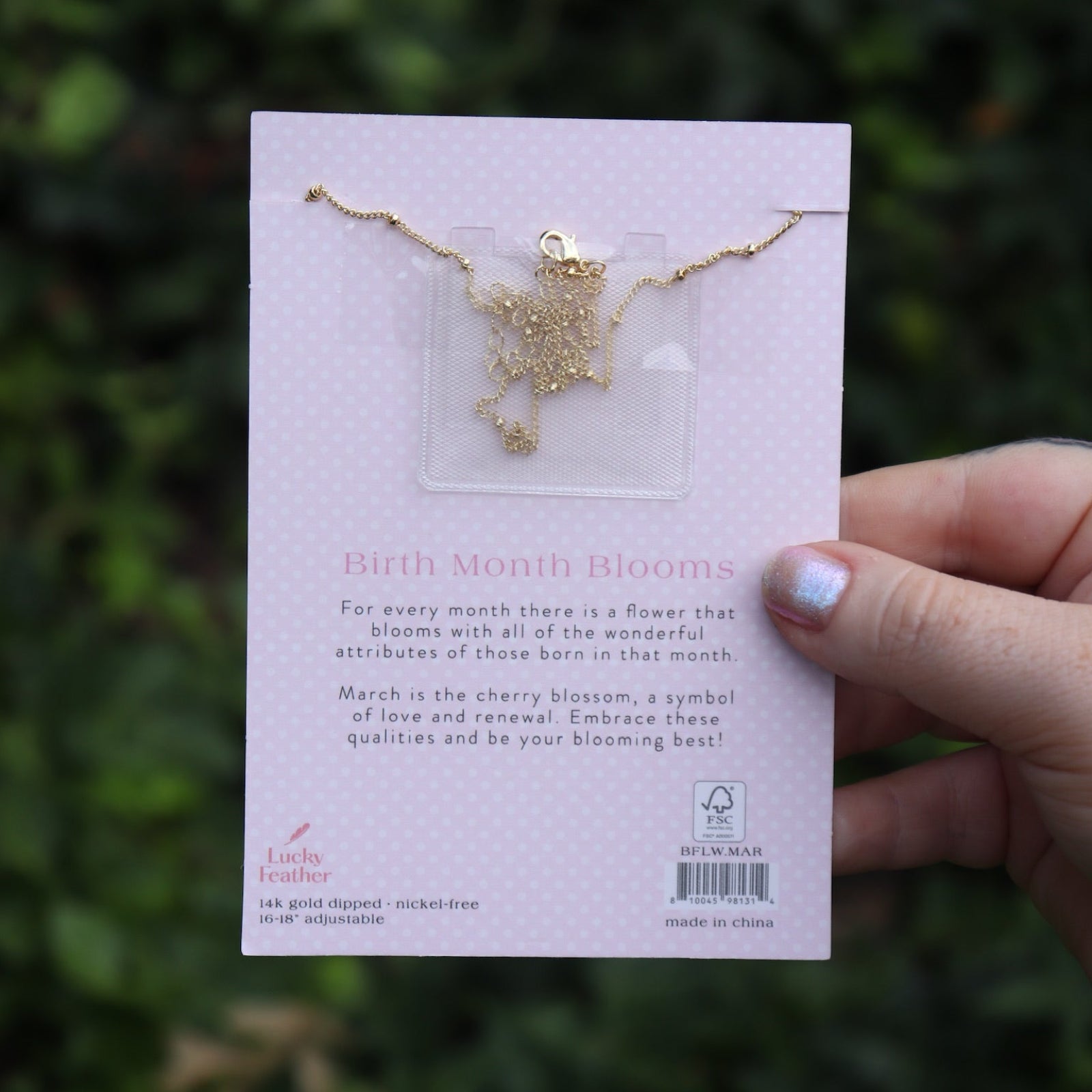 Someone holding the cherry blossom 14k Gold Dipped Nickel-Free Birth Month Flower Necklace in front of greenery showing off the back.