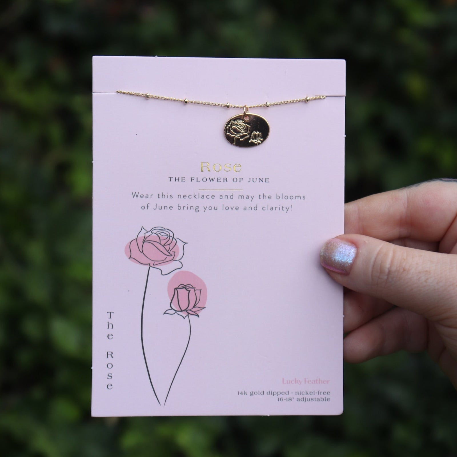 Someone holding the rose 14k Gold Dipped Nickel-Free Birth Month Flower Necklace in front of greenery.