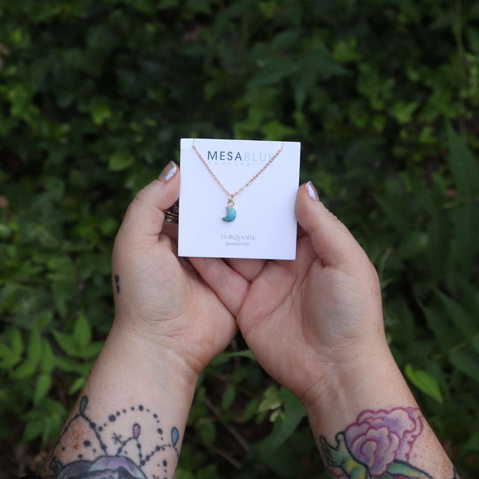 Someone holding the Tiny Turquoise Moon Necklace in front of greenery