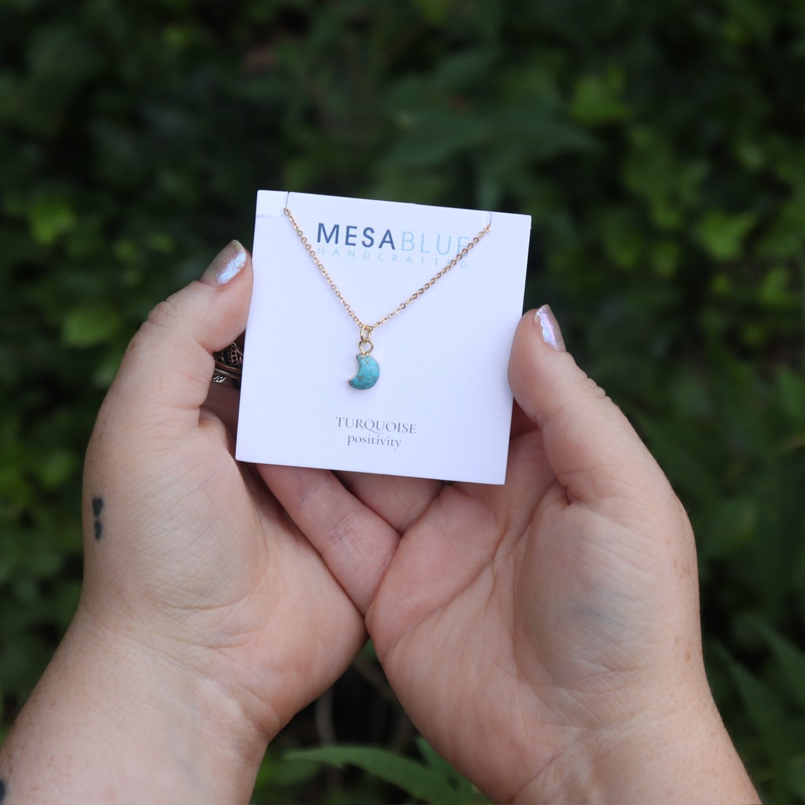 Someone holding the Tiny Turquoise Moon Necklace in front of greenery