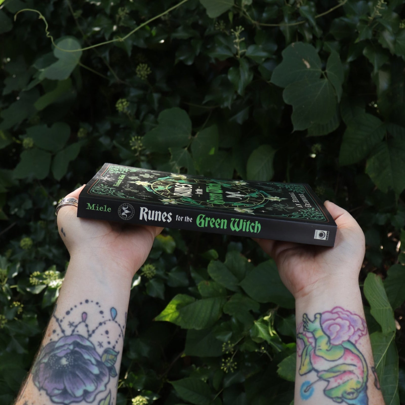 Someone holding Runes for the Green Witch by Nicolette Miele in front of greenery showing off the book's spine.