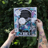 Someone holding the Tarot Coloring - Coloring Book in front of greenery showing the back.