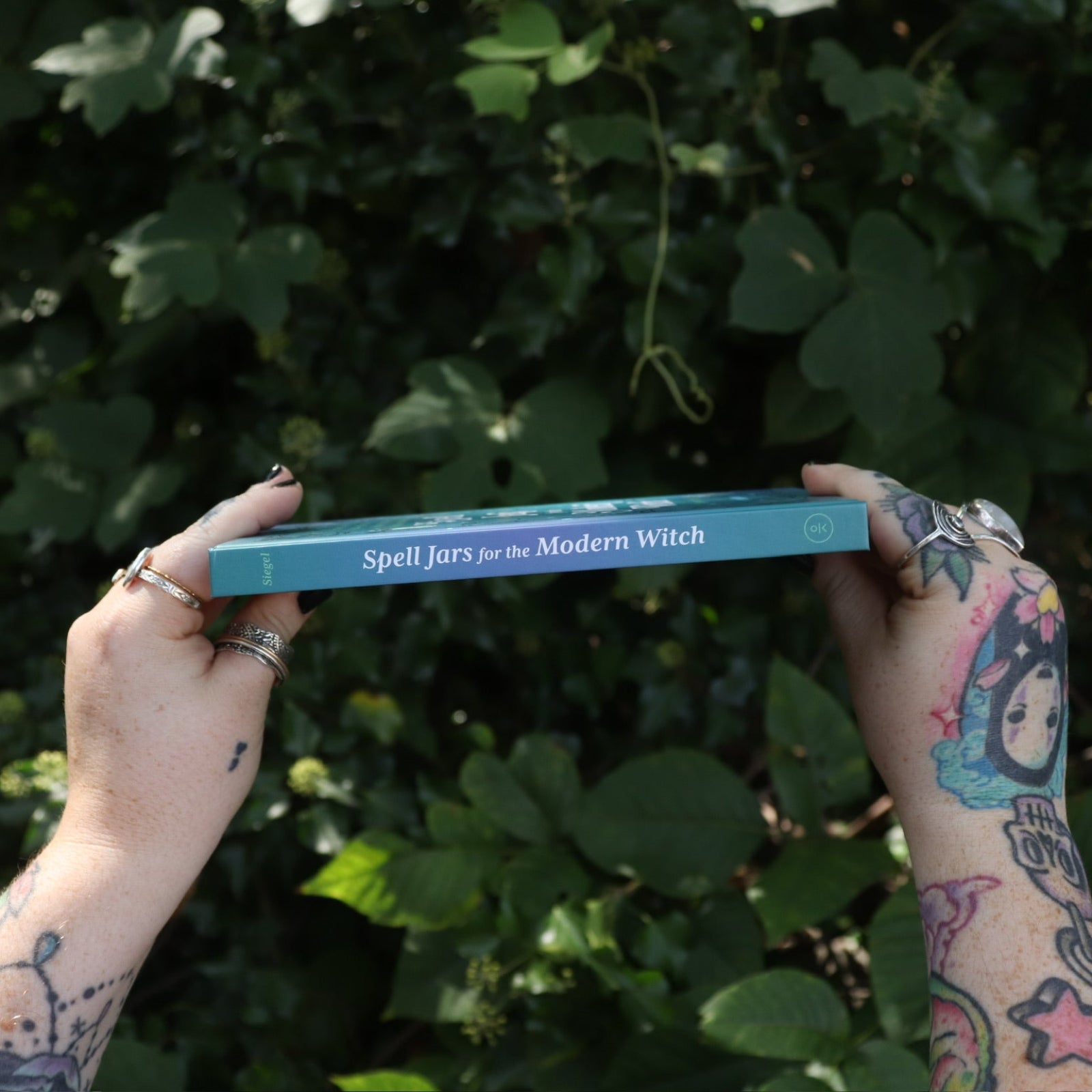 Someone holding the Spell Jars for the Modern Witch Book in front of greenery, showing the spine of the book.
