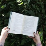 Someone holding the Green Witch by Arin Murphy-Hiscock open in front of greenery, showing off the pages.