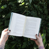 Someone holding the Green Witch by Arin Murphy-Hiscock open in front of greenery, showing off the pages.