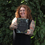 A woman holding the Green Witch by Arin Murphy-Hiscock in front of greenery.