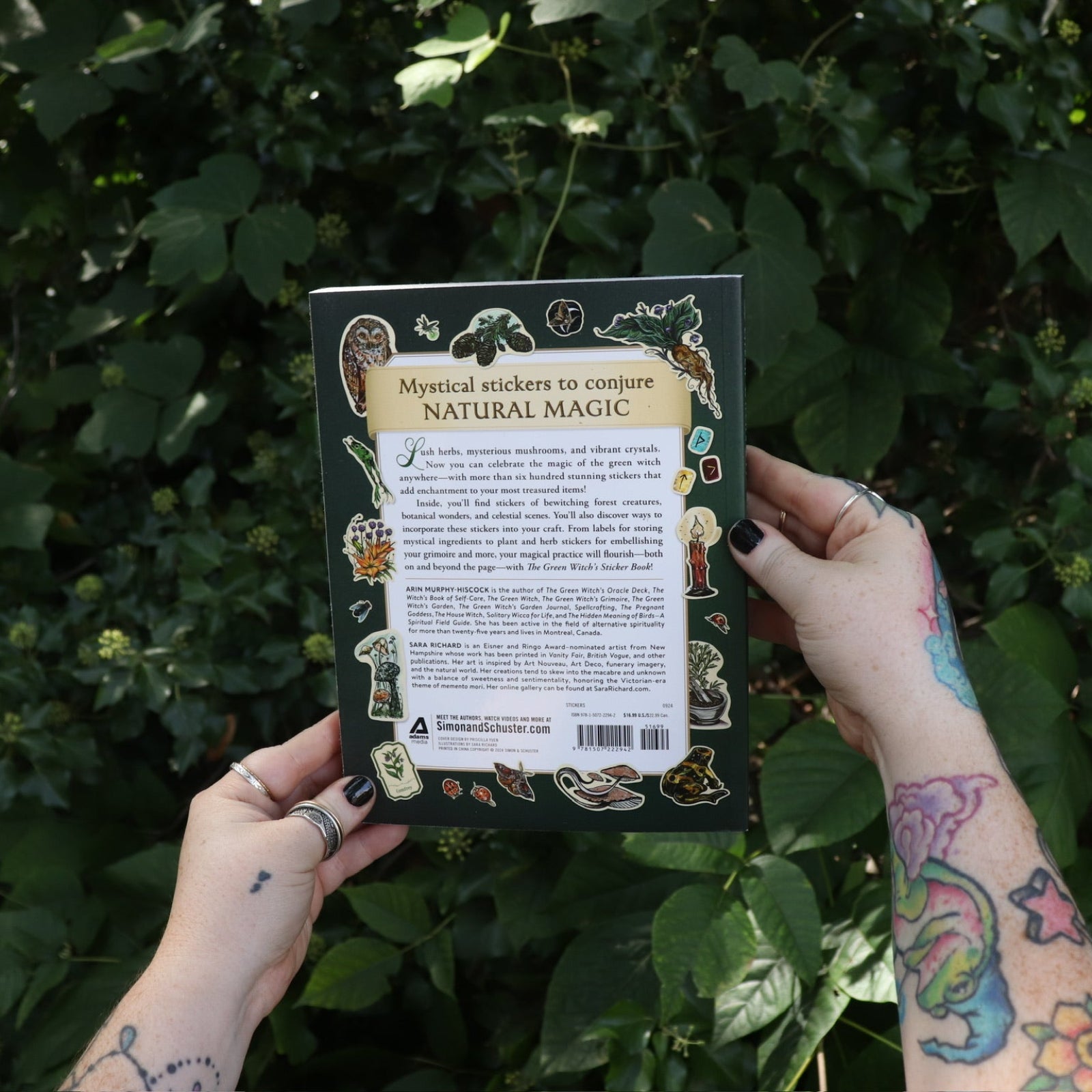 Someone holding the Green Witch's Sticker Book by Arin Murphy-Hiscock in front of greenery, showing off the back of the book.