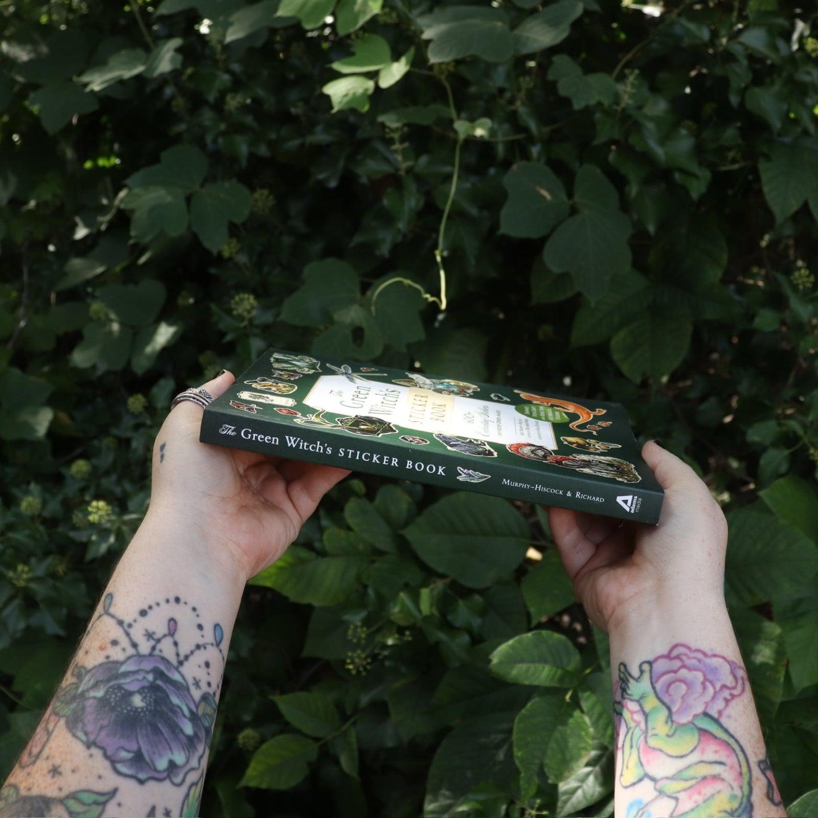 Someone holding the Green Witch's Sticker Book by Arin Murphy-Hiscock in front of greenery, showing off the spine of the book.