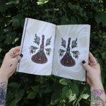 Someone holding the Green Witch's Sticker Book by Arin Murphy-Hiscock open in front of greenery, showing odd the sticker pages.