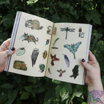 Someone holding the Green Witch's Sticker Book by Arin Murphy-Hiscock open in front of greenery, showing odd the sticker pages.