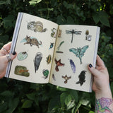 Someone holding the Green Witch's Sticker Book by Arin Murphy-Hiscock open in front of greenery, showing odd the sticker pages.