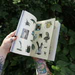 Someone holding the Green Witch's Sticker Book by Arin Murphy-Hiscock open in front of greenery, showing odd the sticker pages.