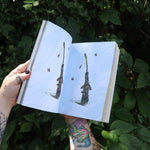Someone holding the Green Witch's Sticker Book by Arin Murphy-Hiscock open in front of greenery, showing odd the sticker pages.