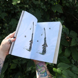 Someone holding the Green Witch's Sticker Book by Arin Murphy-Hiscock open in front of greenery, showing odd the sticker pages.