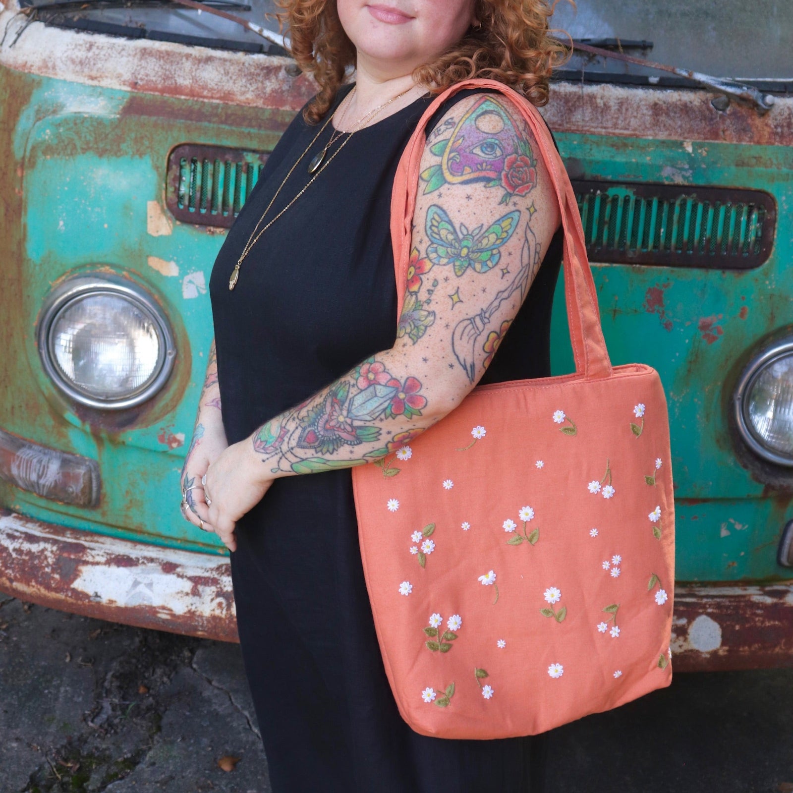 A woman has the Daisy Embroidered Orange Linen Zipper Tote Bag on her shoulder while wearing a black tank dress.