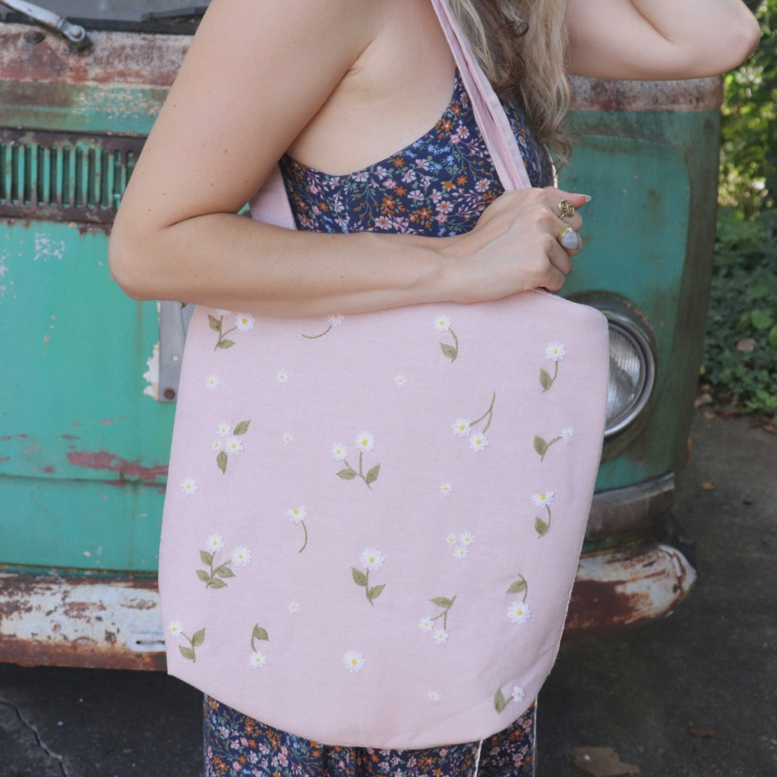 A woman wearing a navy floral dress has the Daisy Embroidered Pastel Pink Linen Zipper Tote Bag on her shoulder.