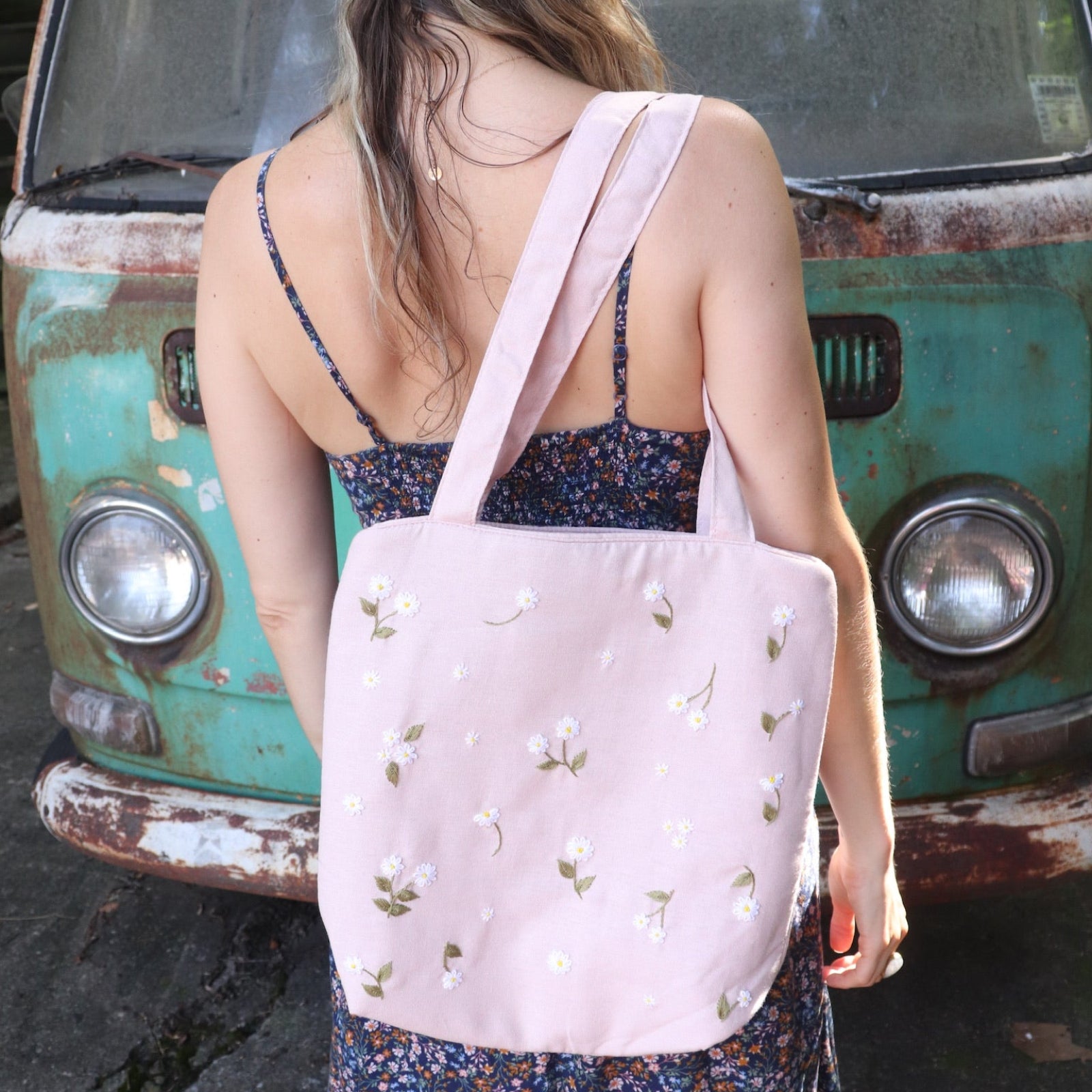 A woman wearing a navy floral dress has the Daisy Embroidered Pastel Pink Linen Zipper Tote Bag on her shoulder.