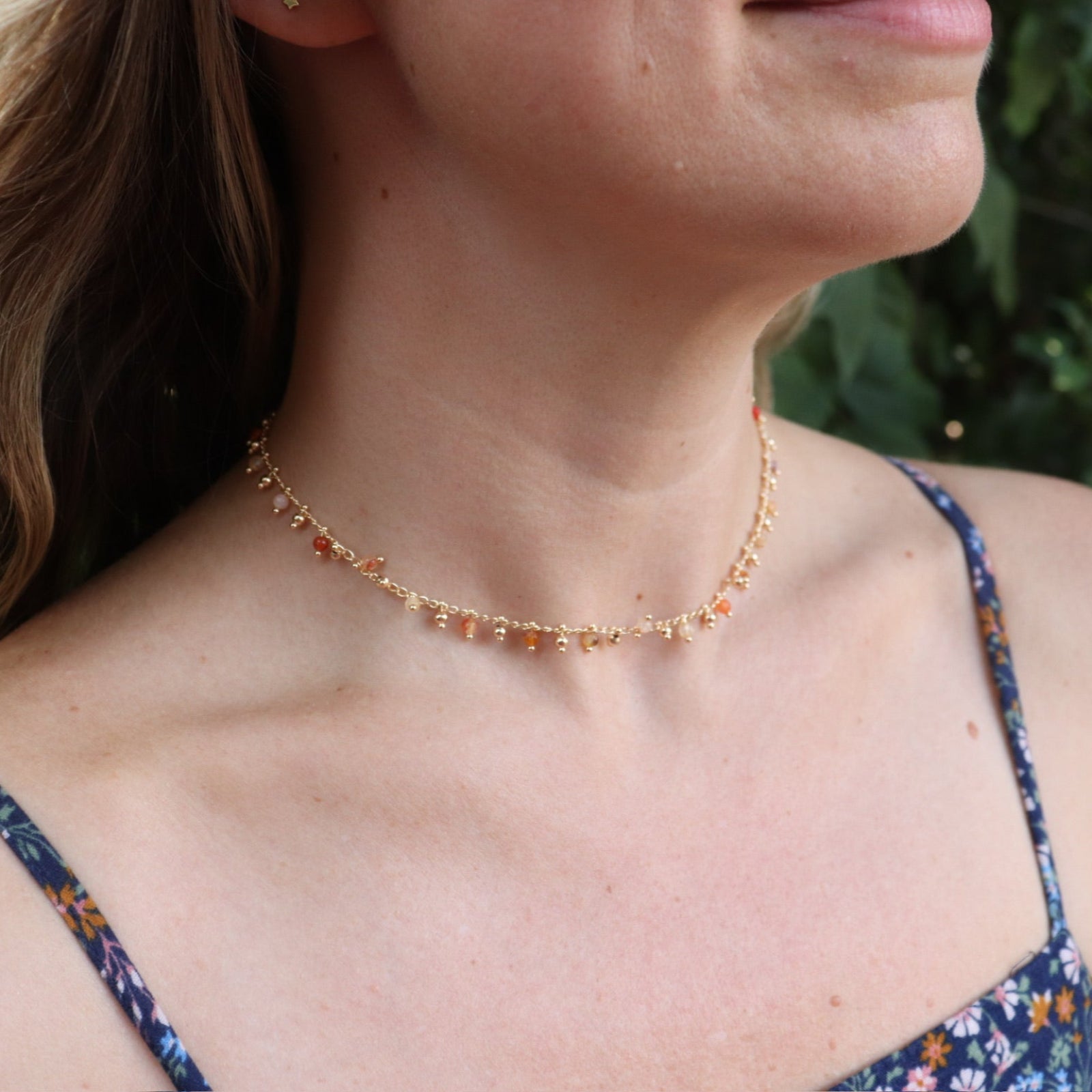 A woman wearing the orange Midsummer Gemstone Agate Gold Plated Necklace in front of some greenery.