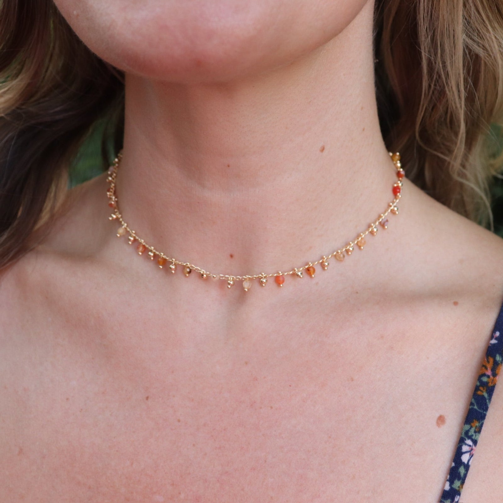 A woman wearing the orange Midsummer Gemstone Agate Gold Plated Necklace in front of some greenery.