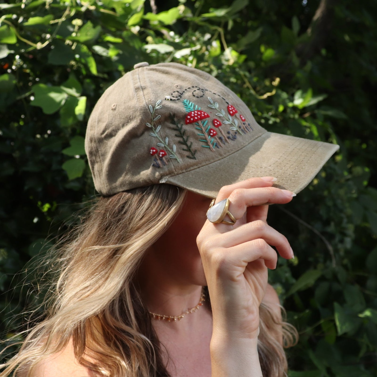 A woman wearing the Mushroom Forest & Bee Embroidered Adjustable Baseball Cap in front of greenery.