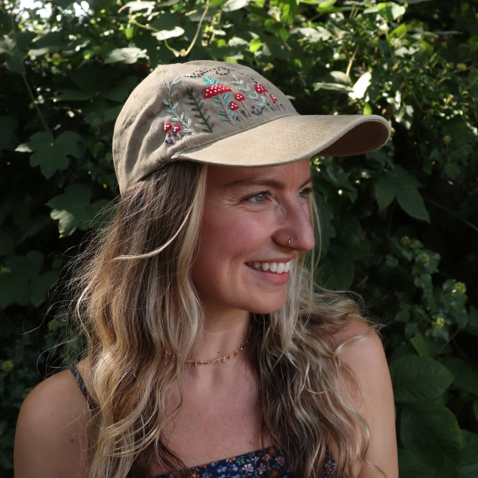 A woman wearing the Mushroom Forest & Bee Embroidered Adjustable Baseball Cap with a navy floral dress in front of greenery.