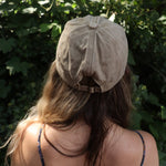 A woman wearing the Mushroom Forest & Bee Embroidered Adjustable Baseball Cap with a navy floral dress in front of greenery, showing the back.