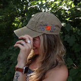 A woman wearing the Orange Pansy Embroidered Adjustable Baseball Cap with a navy floral dress in front of the greenery.