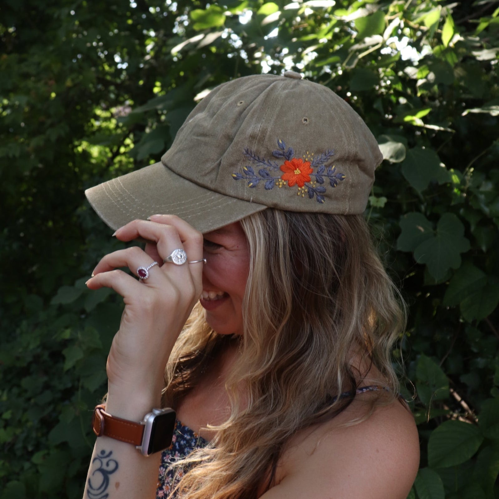 A woman wearing the Orange Pansy Embroidered Adjustable Baseball Cap with a navy floral dress in front of the greenery.