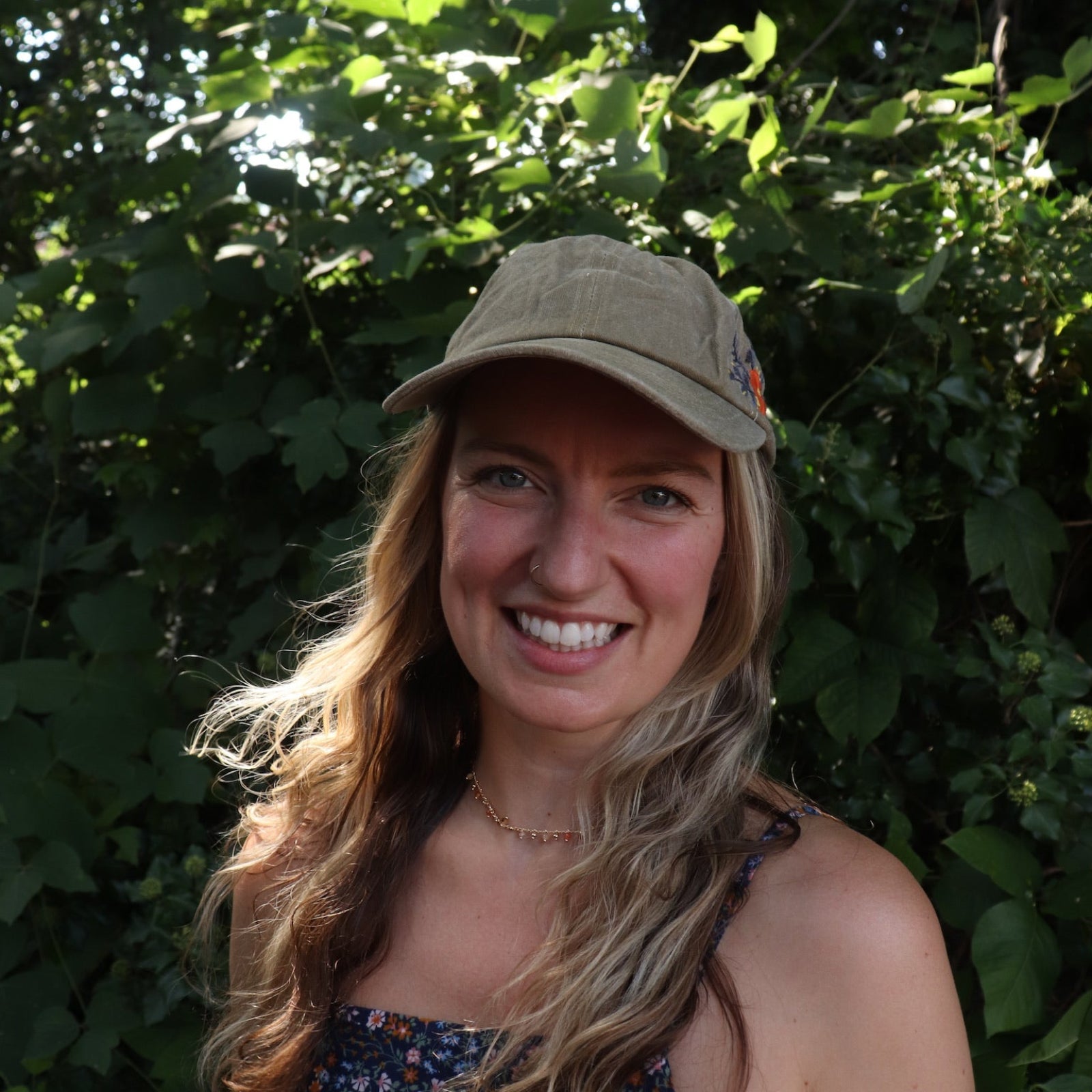A woman wearing the Orange Pansy Embroidered Adjustable Baseball Cap with a navy floral dress in front of the greenery.