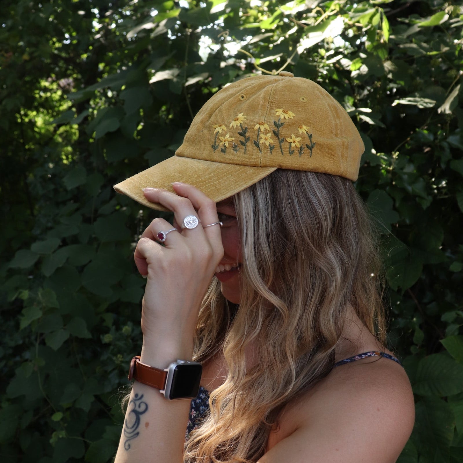 A woman wearing the Daisy Garden Embroidered Adjustable Baseball Cap with a navy floral dress in front of greenery.