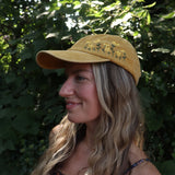 A woman wearing the Daisy Garden Embroidered Adjustable Baseball Cap with a navy floral dress in front of greenery.