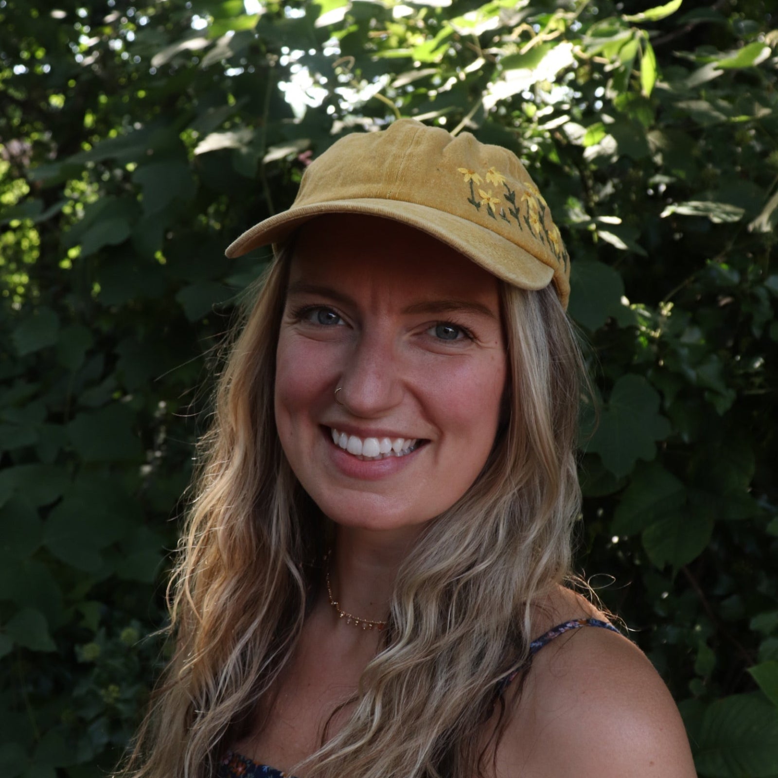 A woman wearing the Daisy Garden Embroidered Adjustable Baseball Cap with a navy floral dress in front of greenery.