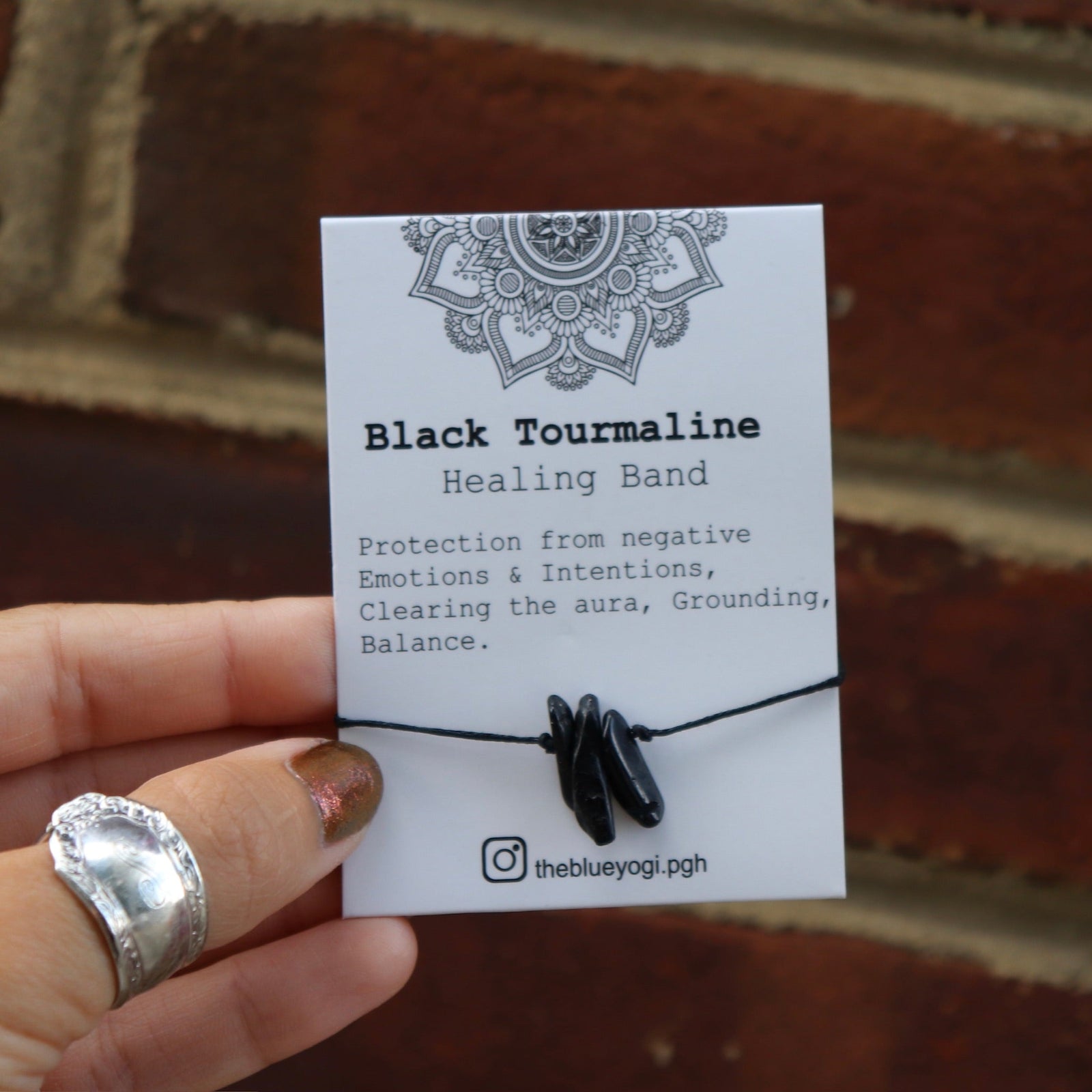 Someone holding the Black Tourmaline Healing Adjustable Band with an Affirmation in front of a brick wall.