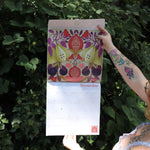 Someone holding the Nina Pace Wall Calendar 2026 open in front of greenery, showing off the pages.
