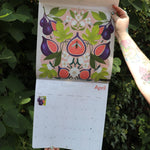 Someone holding the Nina Pace Wall Calendar 2026 open in front of greenery, showing off the pages.