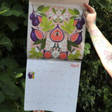 Someone holding the Nina Pace Wall Calendar 2026 open in front of greenery, showing off the pages.