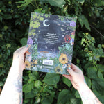 Someone holding the Green Witch Illustrated by Arin Murphy-Hiscock in front of the greenery, showing off the back.
