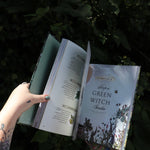 Someone holding the Green Witch Illustrated by Arin Murphy-Hiscock open in front of the greenery, showing off the pages.