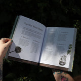 Someone holding the Green Witch Illustrated by Arin Murphy-Hiscock open in front of the greenery, showing off the pages.