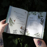 Someone holding the Green Witch Illustrated by Arin Murphy-Hiscock open in front of the greenery, showing off the pages.