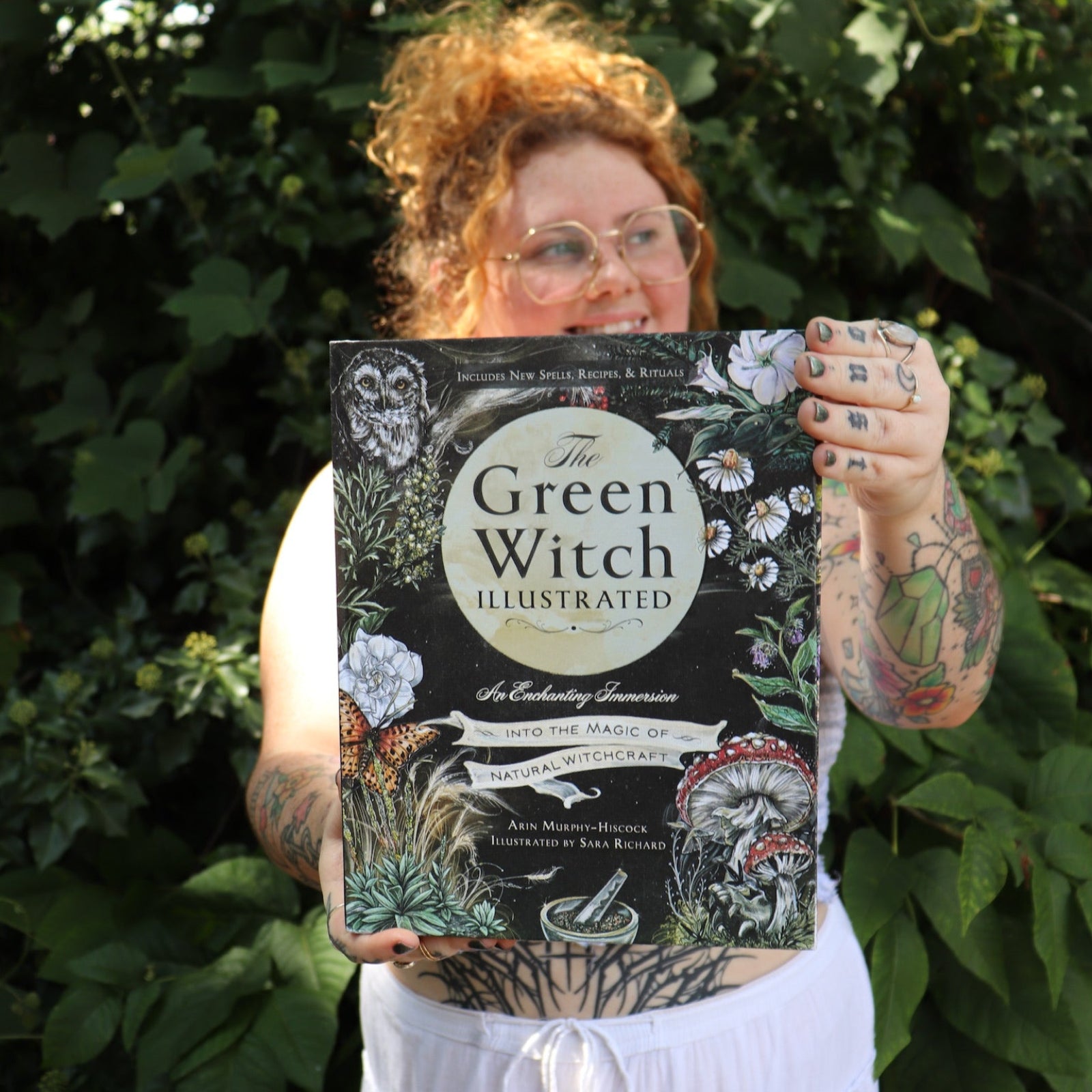 A woman holding the Green Witch Illustrated by Arin Murphy-Hiscock in front of greenery.