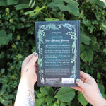 Someone holding the Green Witch's Grimoire by Arin Murphy-Hiscock in front of greenery, showing off the back.