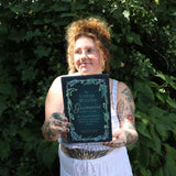 A woman holding the Green Witch's Grimoire by Arin Murphy-Hiscock in front of greenery.