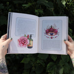 Someone holding the Moon Apothecary by Lorriane  Anderson: Hardcover open in front of greenery, showing off the pages.