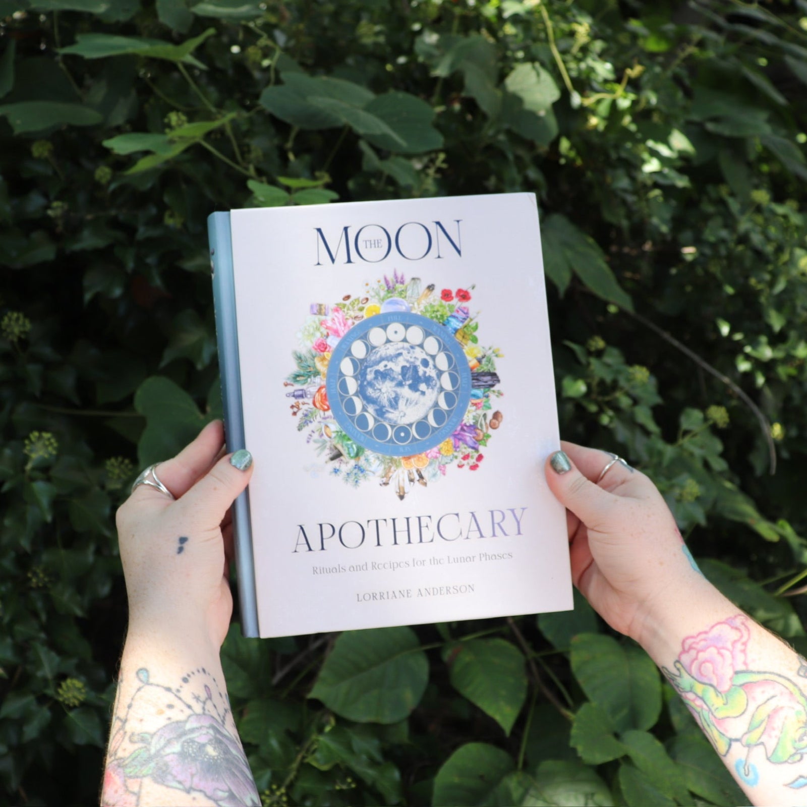 Someone holding the Moon Apothecary by Lorriane  Anderson: Hardcover in front of greenery.
