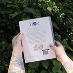 Someone holding the Moon Apothecary by Lorriane  Anderson: Hardcover in front of greenery, showing off the back.