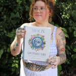 A woman holding the Moon Apothecary by Lorriane  Anderson: Hardcover in front of greenery.