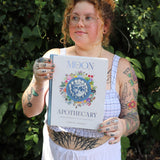A woman holding the Moon Apothecary by Lorriane  Anderson: Hardcover in front of greenery.