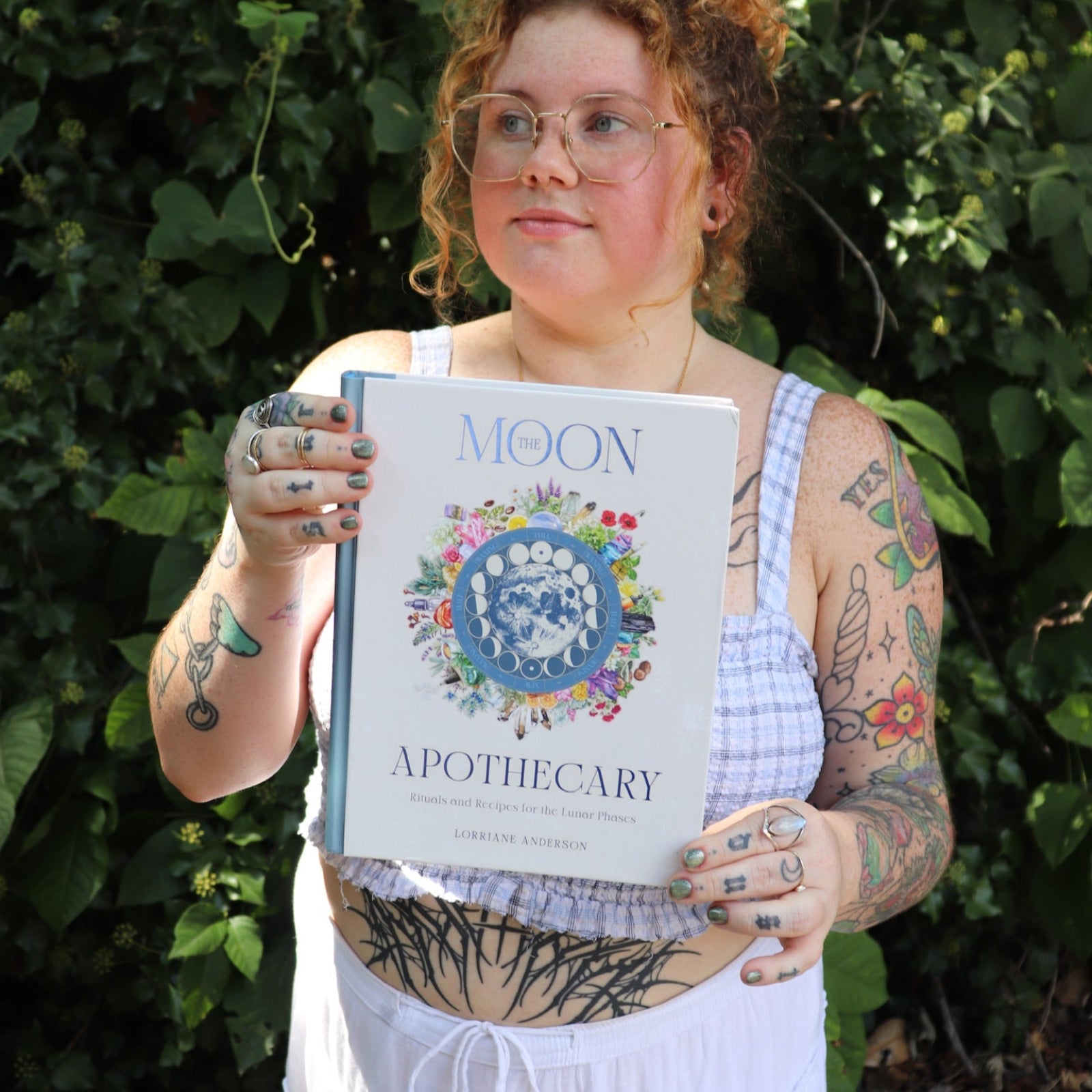 A woman holding the Moon Apothecary by Lorriane  Anderson: Hardcover in front of greenery.